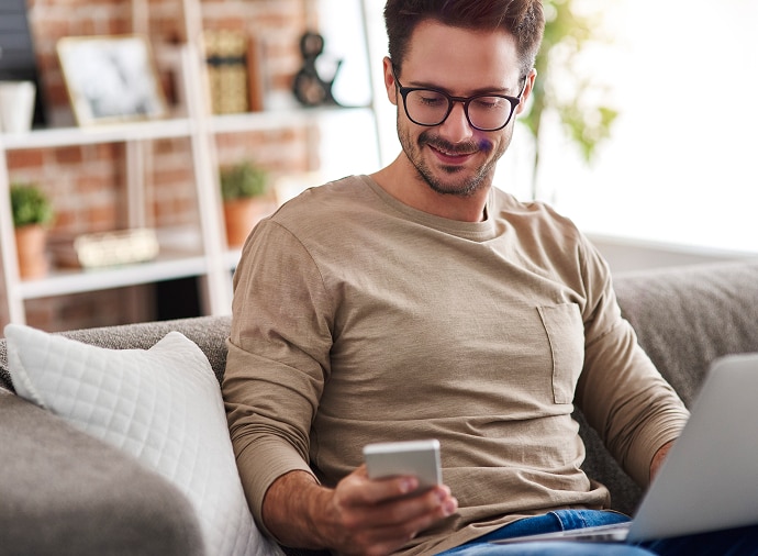 膝の上にノートパソコンを置いて、ソファに座りながらスマートフォンを見る男性